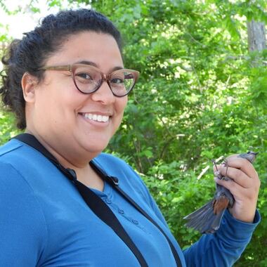 Yamina Nater-Otero, a tan-skinned person with curly hair and glasses, smiles at the camera while holding a Gray Catbird in their left hand. Photo courtesy of Yamina Nater-Otero
